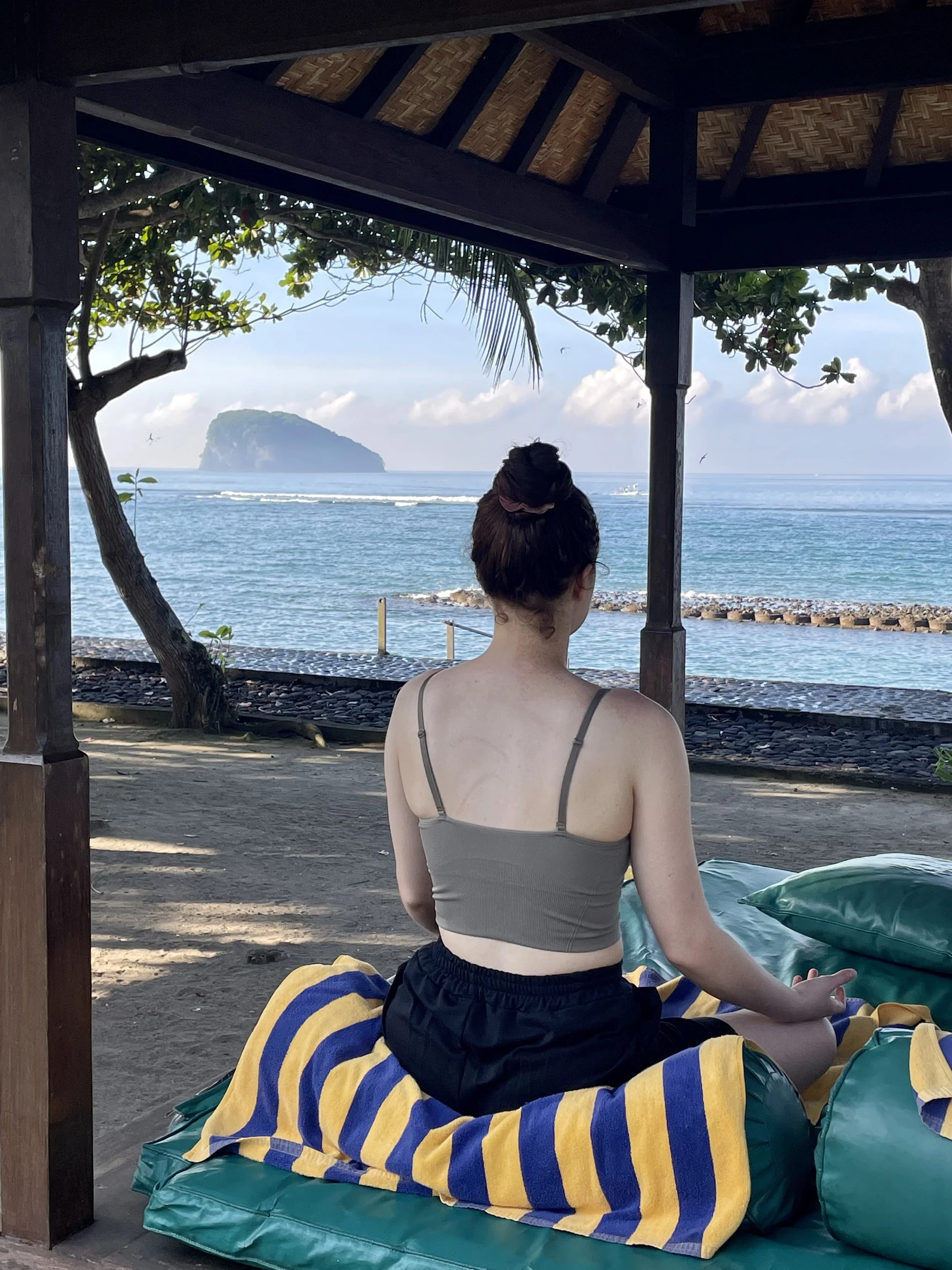 A woman with her hair in a bun, wearing a gray tank top and black shorts, sits cross-legged on a striped yellow and blue towel on a cushioned lounge at a waterfront location, looking at the ocean and a distant island under a partially cloudy sky.