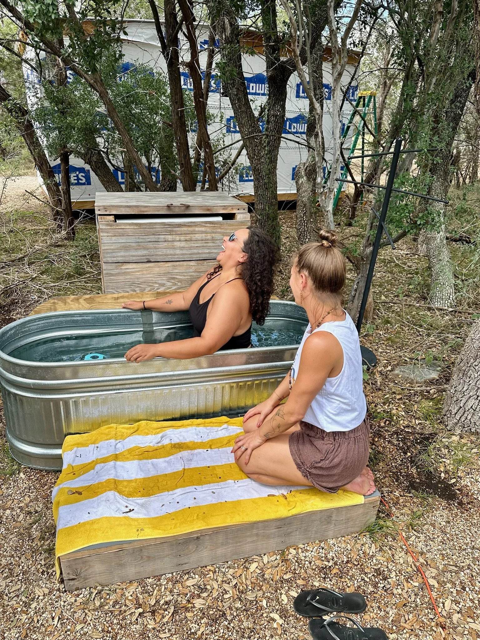 Two women are outdoors near trees, one inside a small metal tub and the other kneeling beside it on a yellow and white striped towel. The woman inside the tub is laughing and wearing sunglasses and a black top. The woman kneeling is dressed in a whit