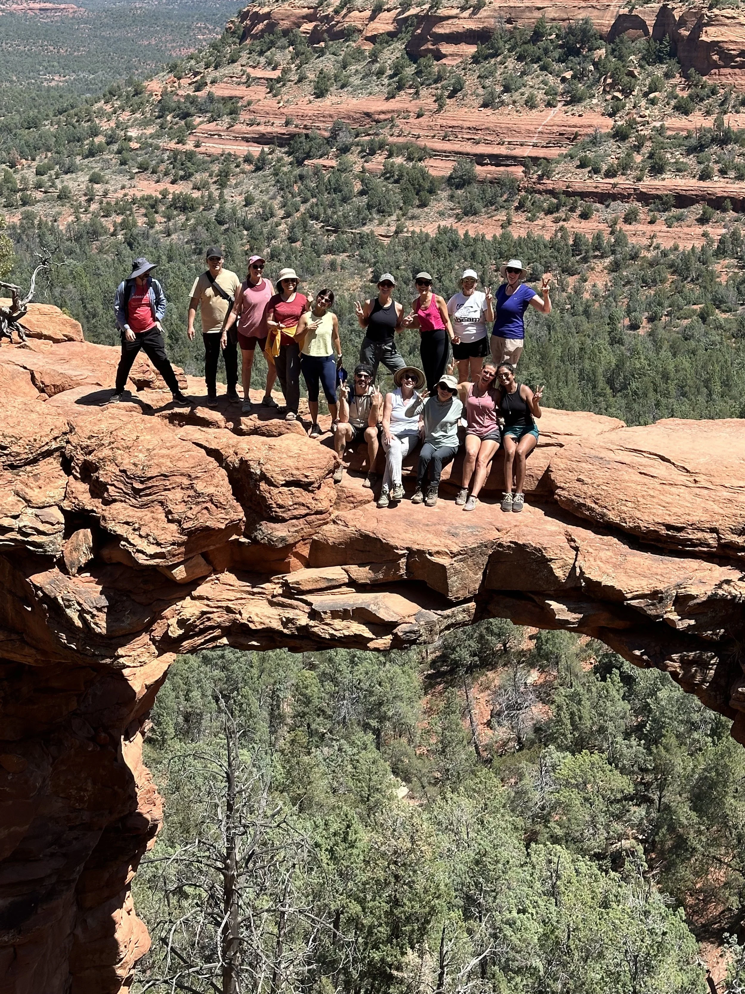 A group of people posing on the red rock formation of Devil's Bridge in Sedona, Arizona, with a natural arch in the foreground and a forested canyon in the background.
