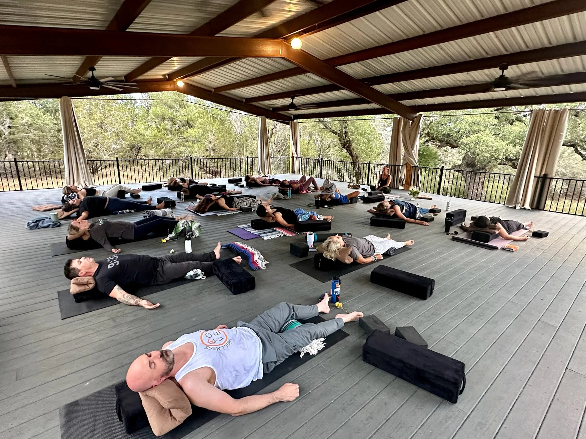 People participating in an outdoor yoga class on a covered deck with trees in the background, lying on yoga mats in a relaxation pose.
