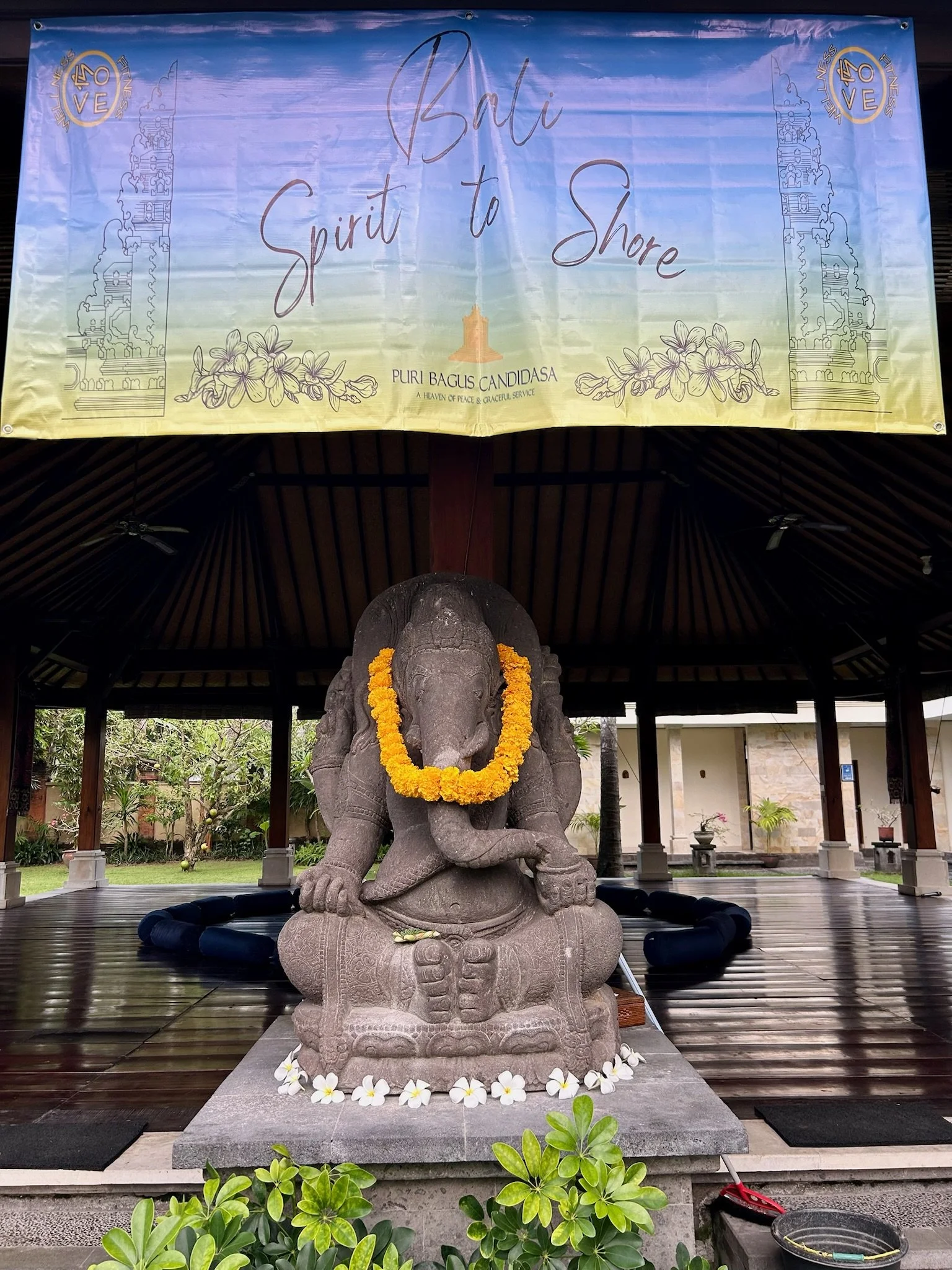 Stone statue of Ganesha with a yellow flower garland, situated under a pavilion with a wooden roof in Bali, Indonesia. The background shows greenery and a large banner that reads 'Bali Spirit to Shore' and features the Puri Bagus Candidasa logo.