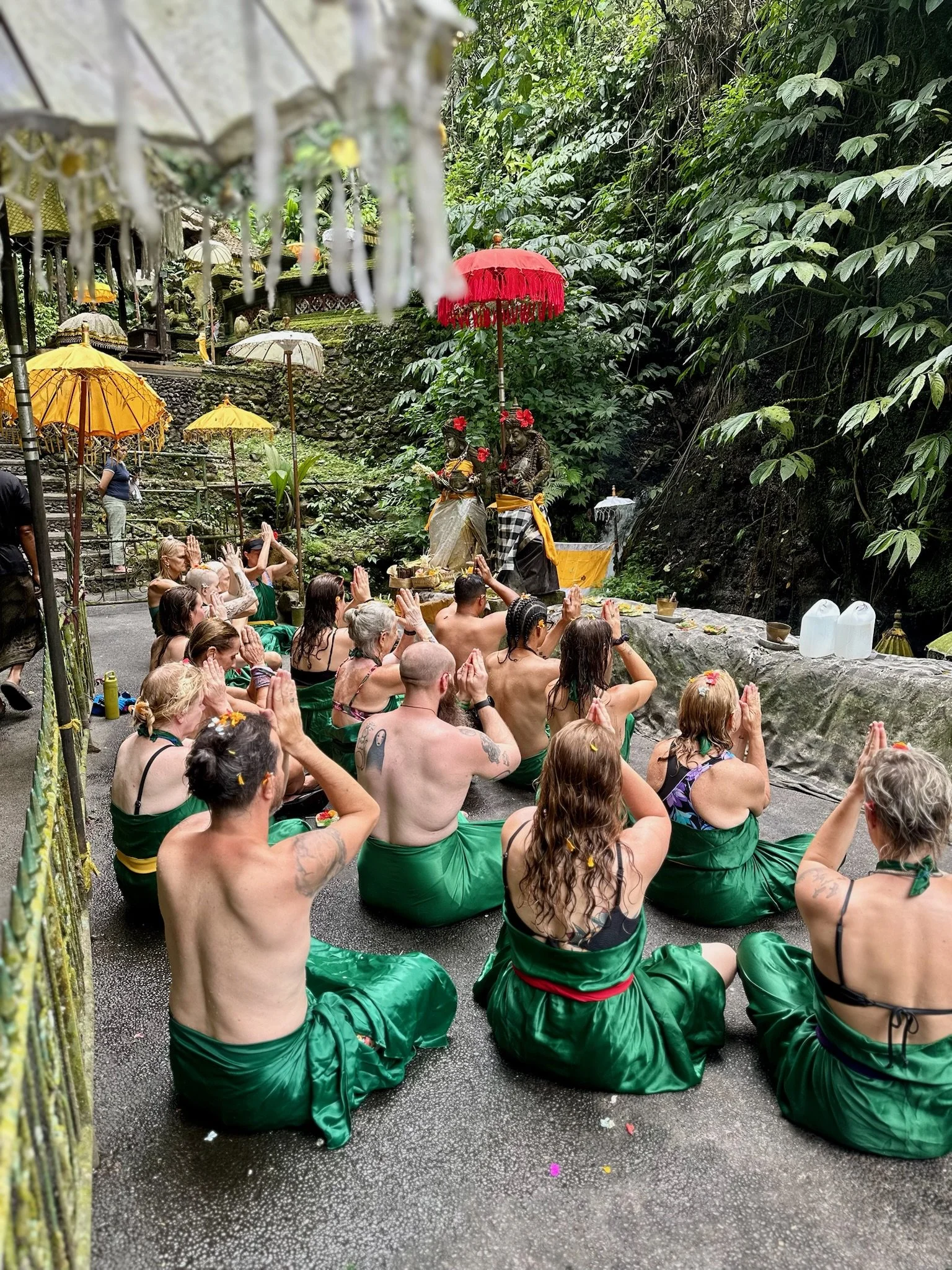 Group of people participating in a traditional Hindu or Balinese prayer ceremony outdoors, seated on the ground with hands pressed together, surrounded by lush green trees and decorated with umbrellas and statues.