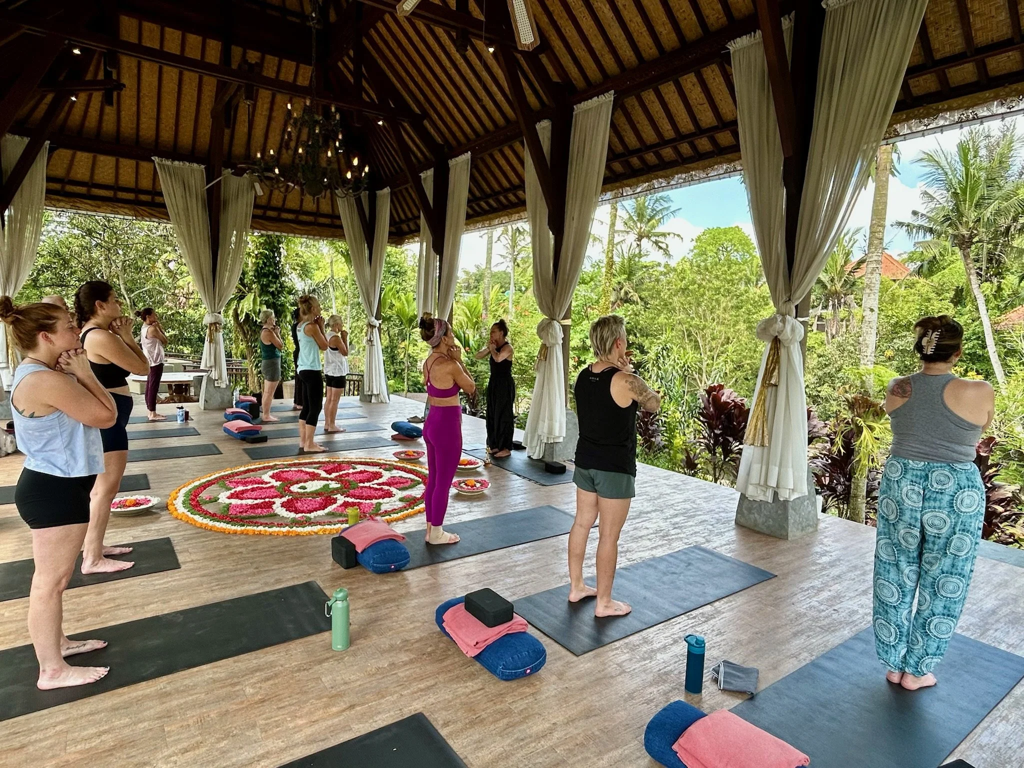 A group of women participating in a yoga class on a covered outdoor deck with draped curtains, surrounded by lush green trees and tropical plants in Bali, Indonesia.