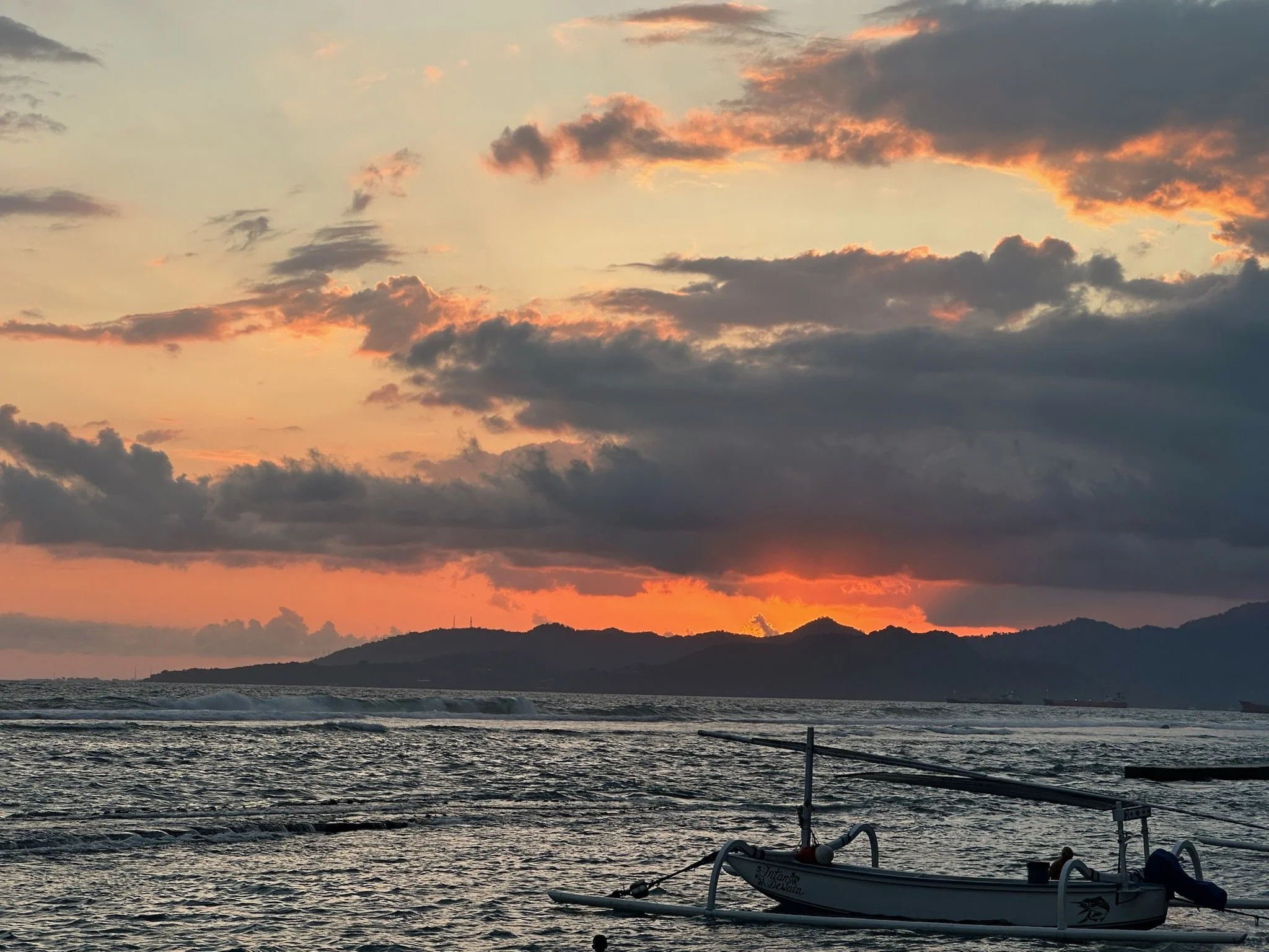 A sunset over the ocean with mountains in the distance and some clouds in the sky. A small boat is in the foreground
