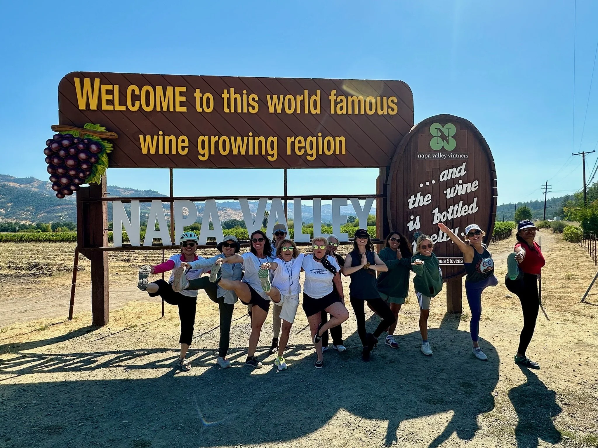Group of women standing with their legs lifted on a sign at Napa Valley vineyards, smiling and posing for the camera on a sunny day.