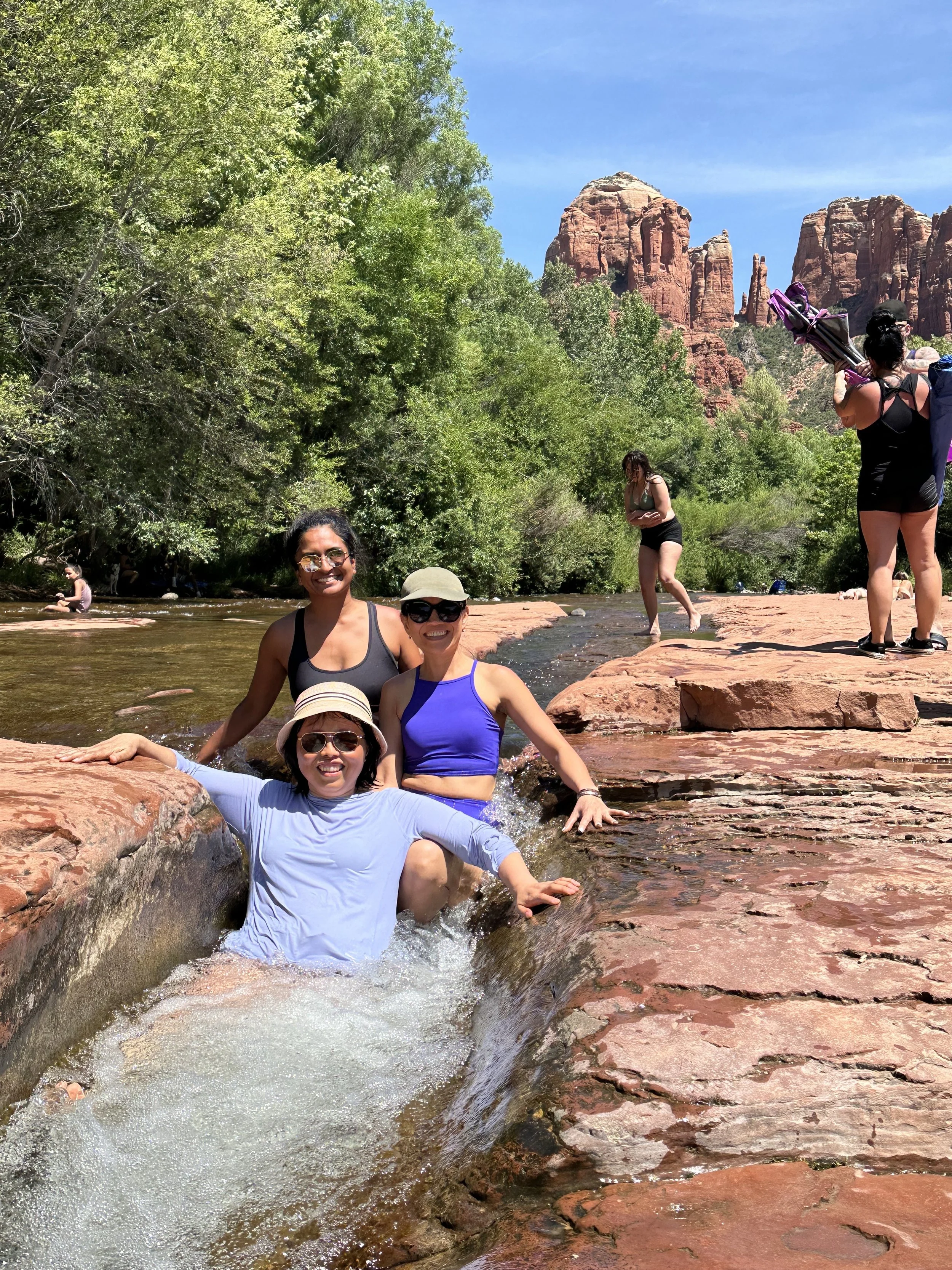 Four women enjoying a shallow stream in a red rock canyon with towering red rock formations and green trees in the background. Two women are sitting in the water, smiling at the camera, while others are standing or walking nearby, some taking photos 
