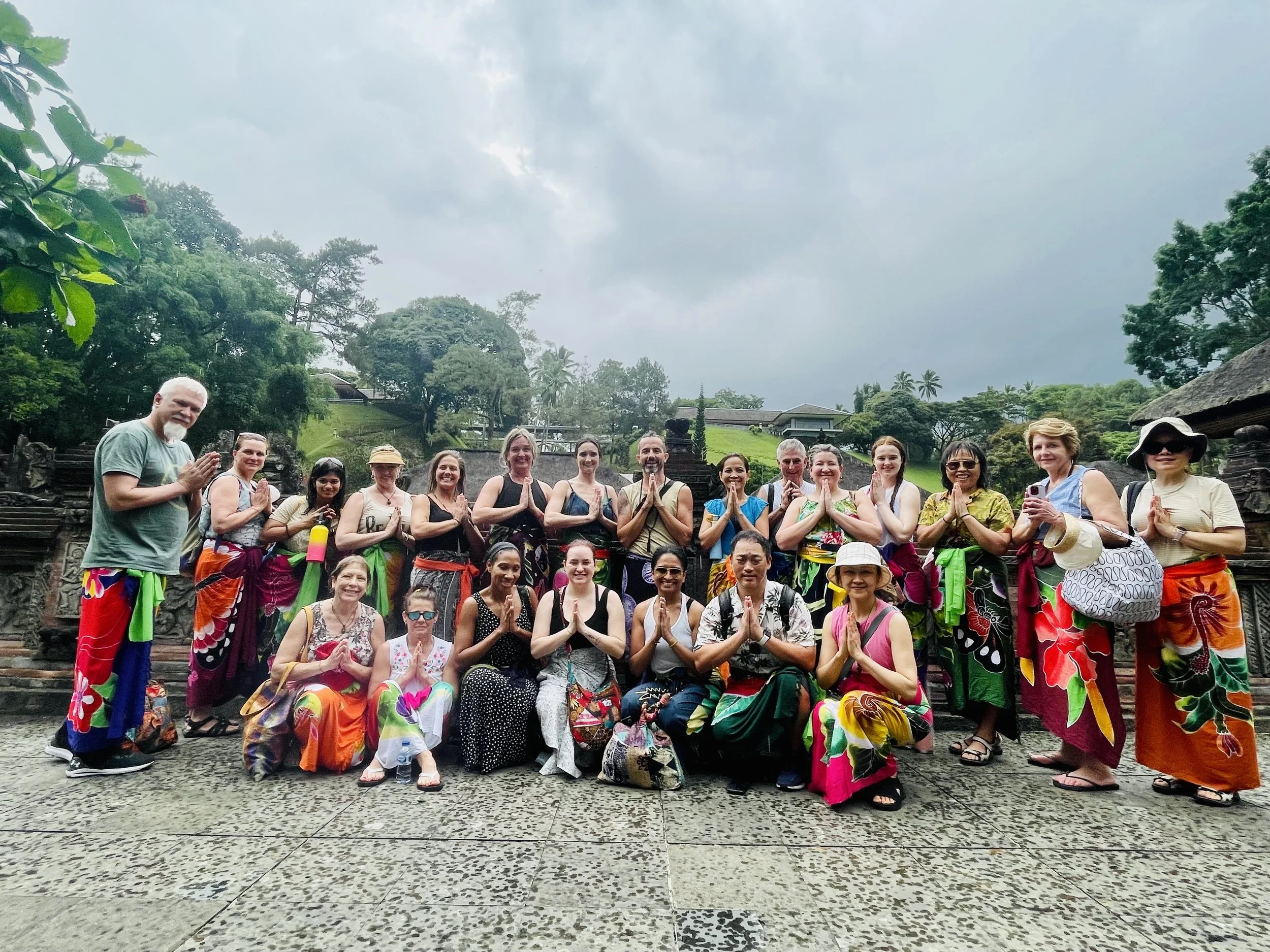 Group of people in colorful traditional attire posing outdoors with hands together in prayer gesture, on cloudy day, lush green background, some taking photos.
