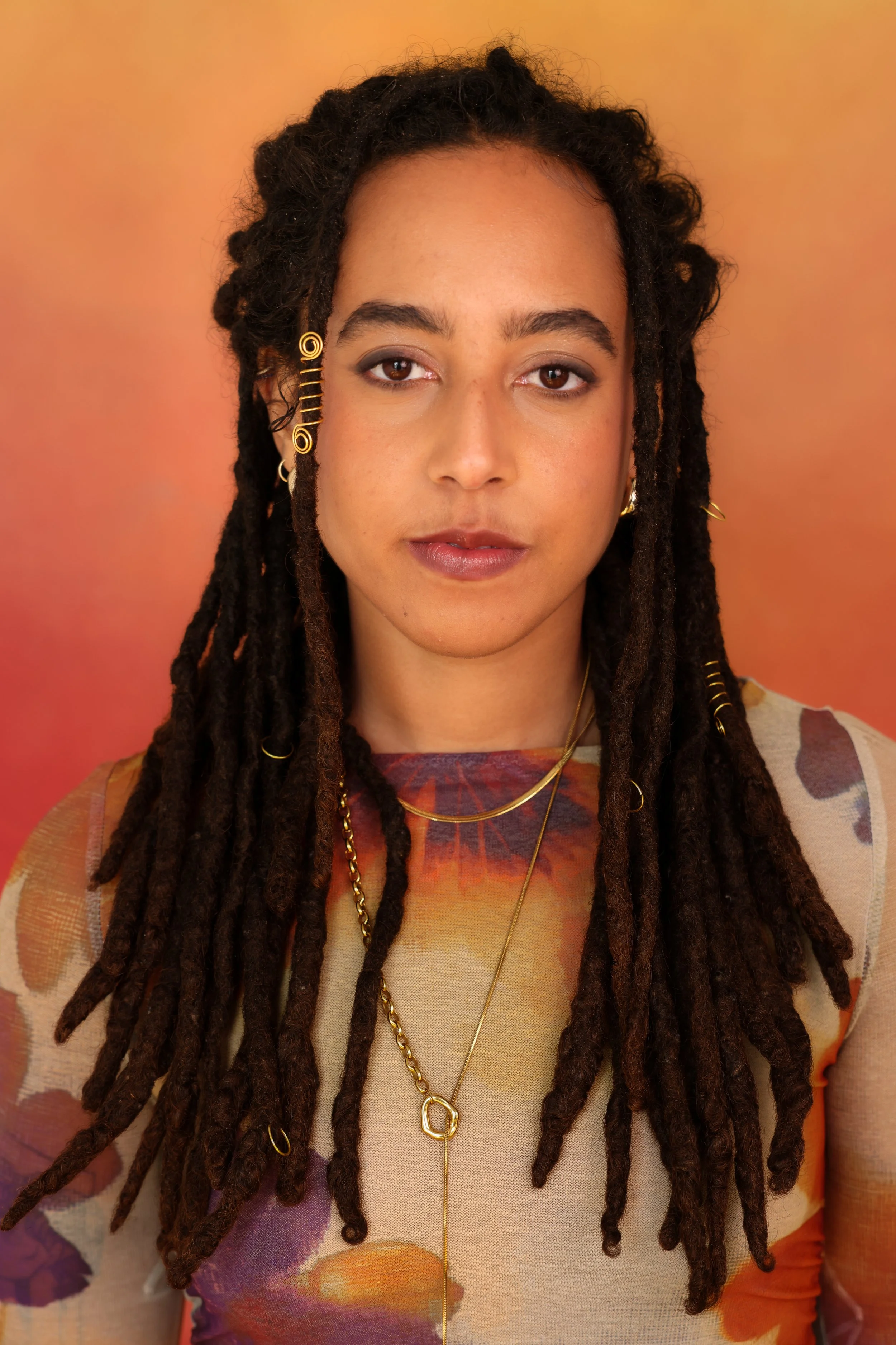 A woman with dreadlocks adorned with gold jewelry and earrings, wearing a sheer floral top, standing against a warm gradient background.