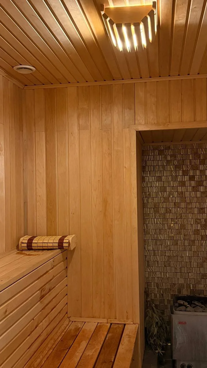 Medinės pirties interjeras su gultais, ant suolo susuktu rankšluosčiu, medine apdaila ir kampe esančia krosnele su akmenimis.