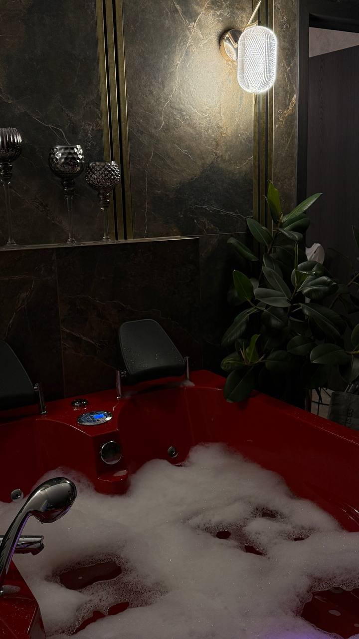Prabangus vonios kambario kampas su raudona sūkurine vonia, pilna putojančio vandens, tamsaus marmuro siena su aukso detalėmis, moderniu sieniniu šviestuvu, dekoratyvinėmis vazomis ir dideliu žaliu augalu.