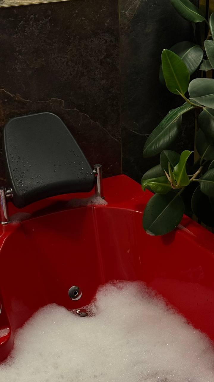 Vonia, pripildyta muilo putų, su pritvirtinta juoda pagalvėle galvai ir šalia esančiu žaliu augalu.