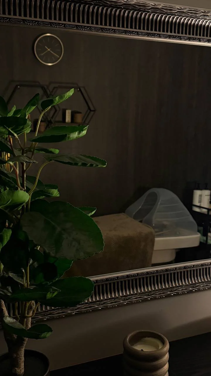 Kambario su tamsaus medžio siena, laikrodžiu, augalu ir vystymo stalu atspindys rėmintame veidrodyje.