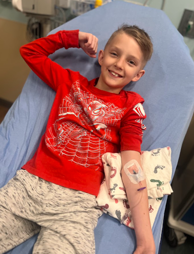 A smiling boy lying on a hospital bed with a bandaged arm, flexing his right arm to show his strength, wearing a red Spider-Man shirt and light-colored pajama pants.