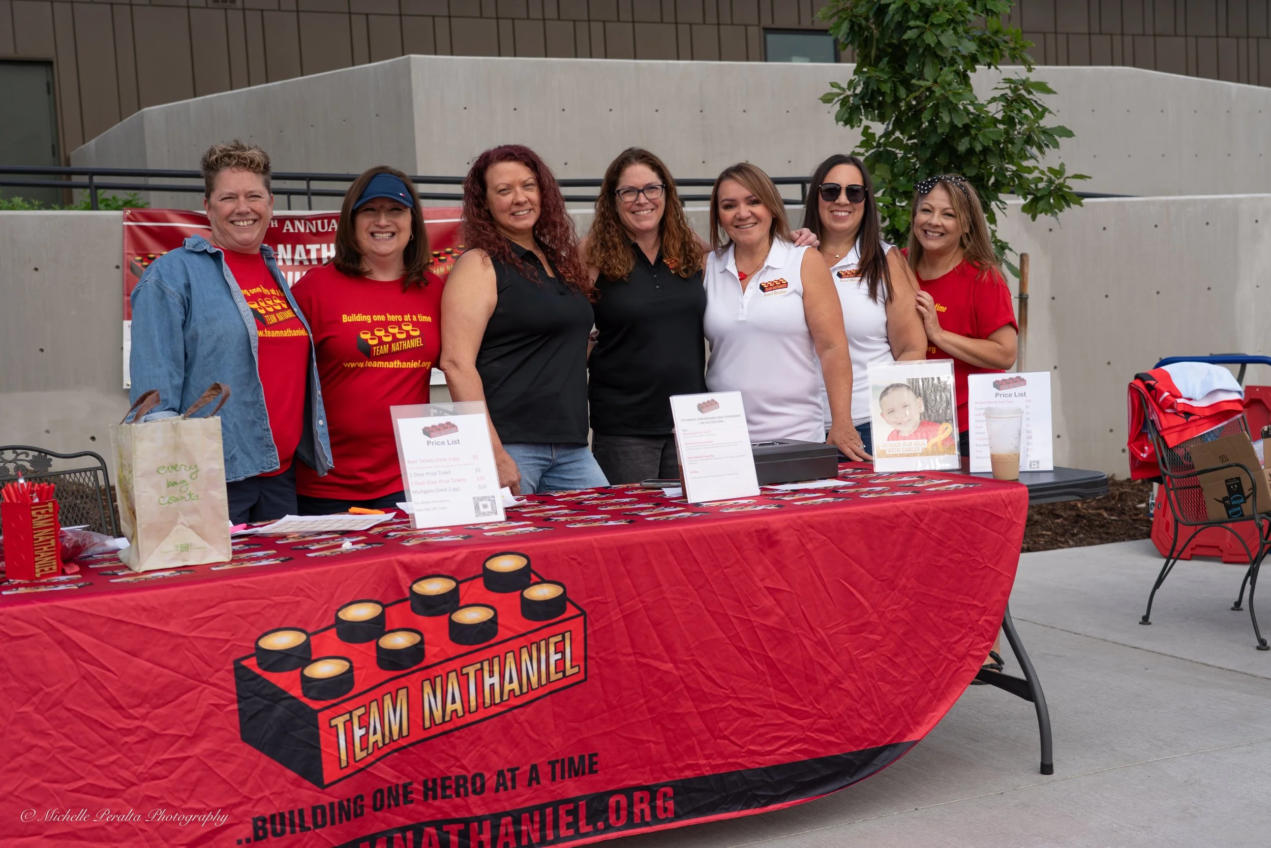Group of seven women standing behind a red event table with Team Nathaniel logo, smiling at the camera, outdoors in front of a building and trees.
