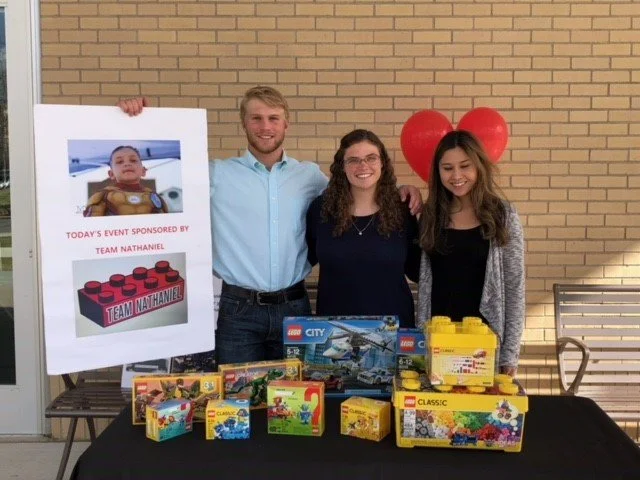Three people smiling at a LEGO event, standing behind a table with LEGO sets and a promotional poster of a child with LEGO bricks, and two red heart-shaped balloons in the background.