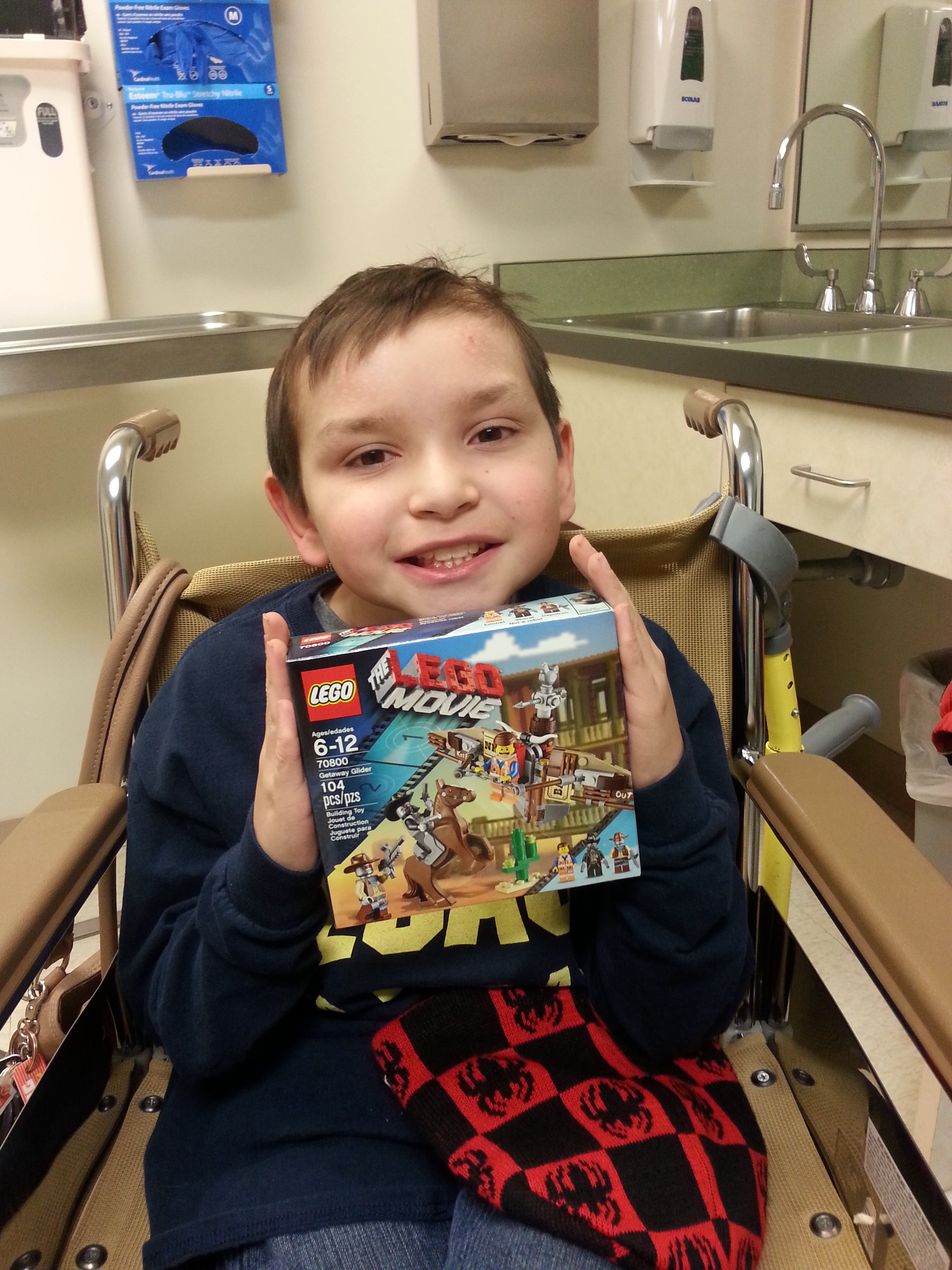A young boy sitting in a wheelchair holding a LEGO The LEGO Movie set, smiling in a medical or hospital room