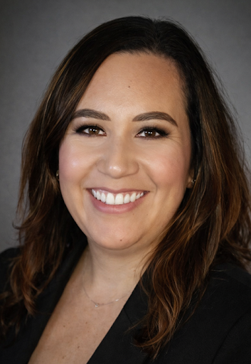 Smiling woman with shoulder-length brown hair, wearing a black blazer, against a gray background.