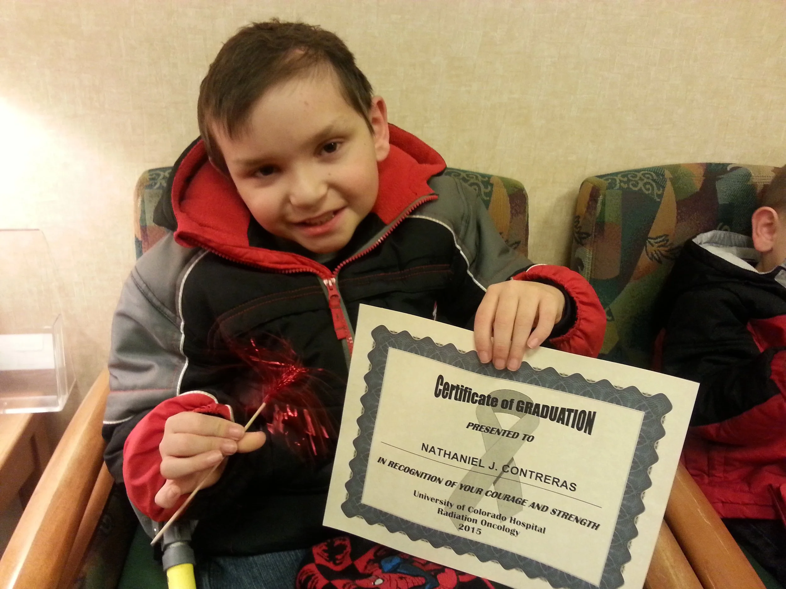 Young boy in a red and black jacket holding a sparkler and a certificate of graduation.