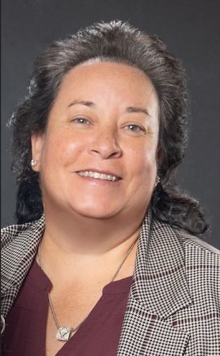 Professional woman with gray hair smiling, wearing a checkered blazer and a maroon top, against a dark background.
