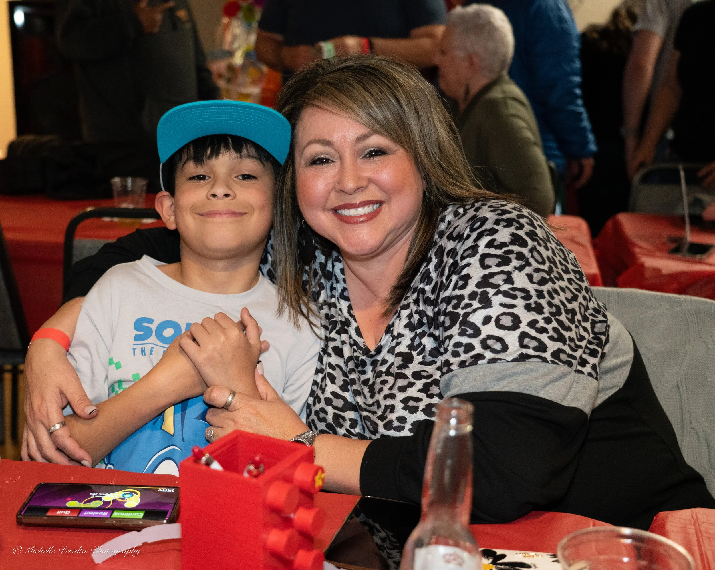 A woman and a boy smiling and embracing each other at a gathering or party, seated at a table with red tablecloths and various items including a phone and a beverage bottle in front of them. The woman is wearing a leopard print top, and the boy is wearing a white T-shirt and a blue cap.