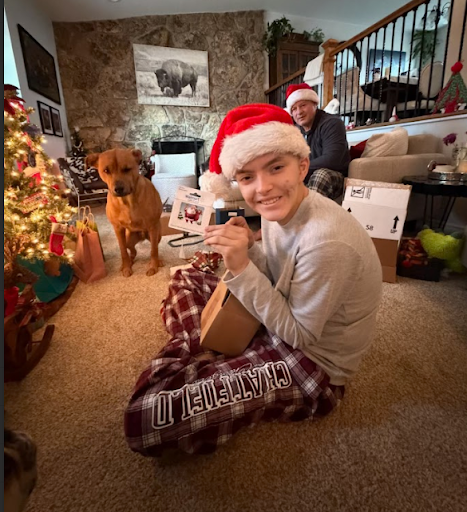 A young boy in pajamas and a Santa hat sitting on the carpet, holding a wrapped gift and smiling at the camera. A dog sits nearby. In the background, an older woman wearing a Santa hat is seated on a sofa with wrapped gifts and holiday decorations around.