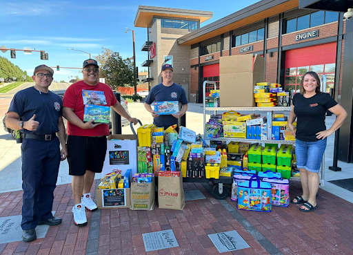 A young girl receiving a LEGO set from a man at a charity event, with two women holding signs promoting the event in the background.