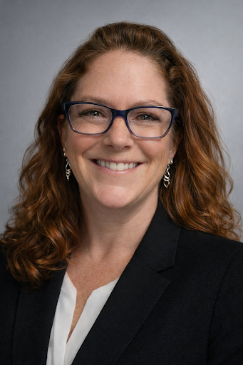 Headshot of a woman with red hair wearing glasses, a black blazer, and a white blouse, smiling at the camera against a gray background.