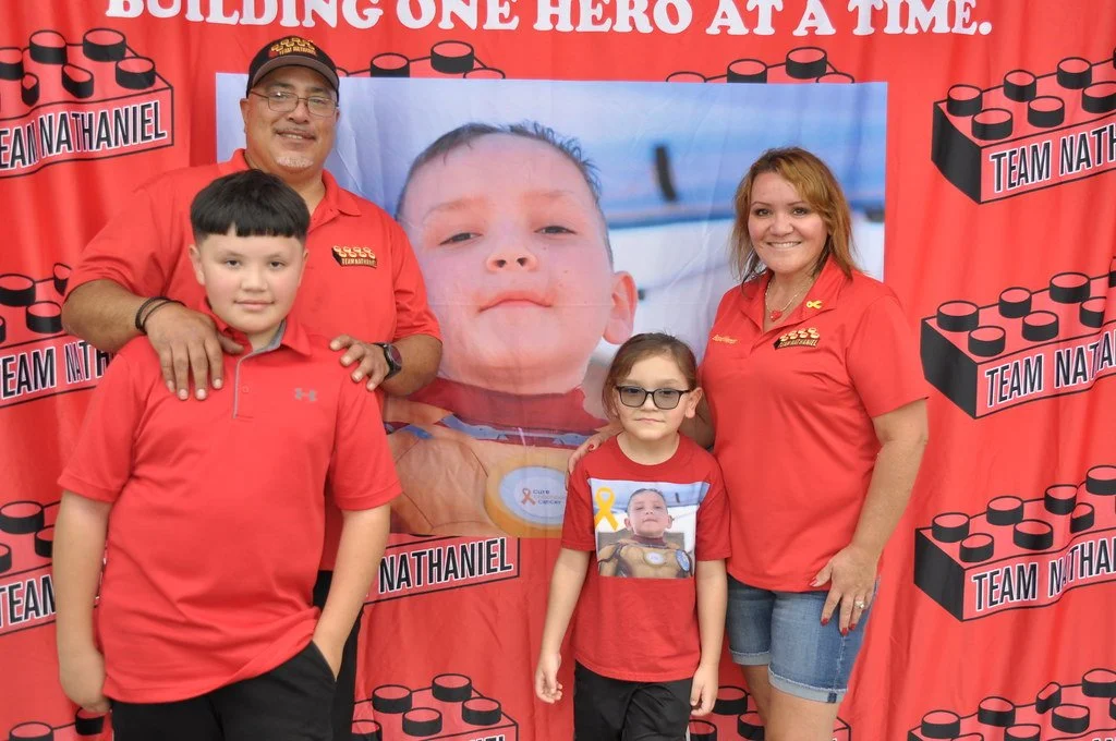 A family of four stands in front of a red backdrop with black block designs and the words "Building one hero at a time" and "Team Nathaniel". They are wearing red shirts, and there is a large photo of a young boy in the background.