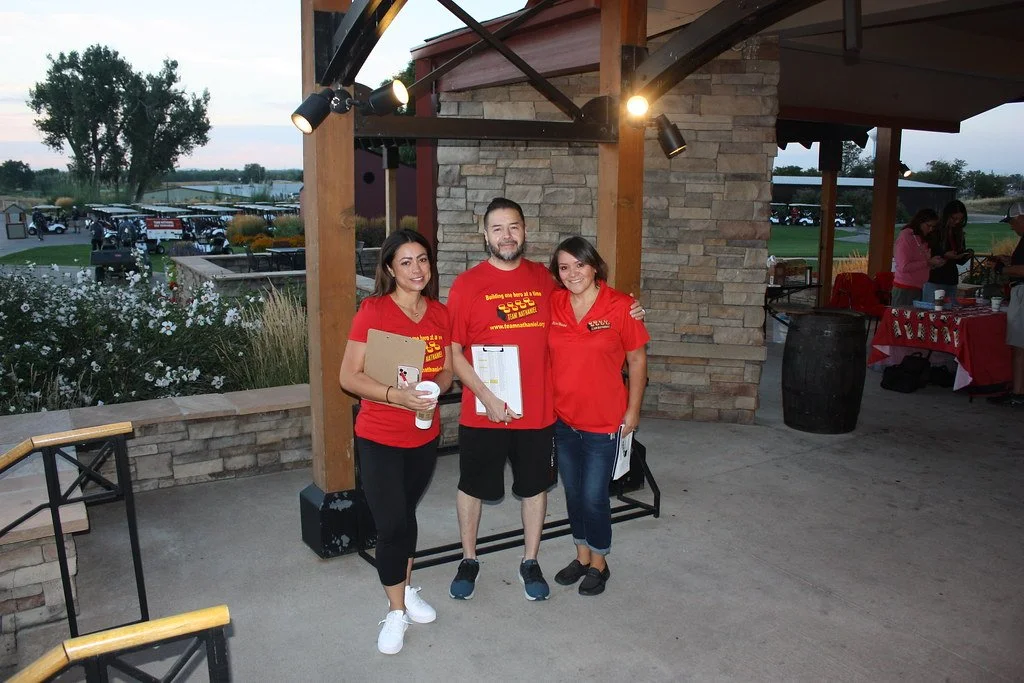 Three smiling people standing outdoors, wearing red shirts, at a gathering or event, with a parking lot and trees in the background.