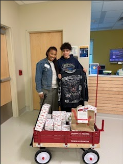 Two children standing next to a red wagon filled with wrapped gifts in a hospital corridor.
