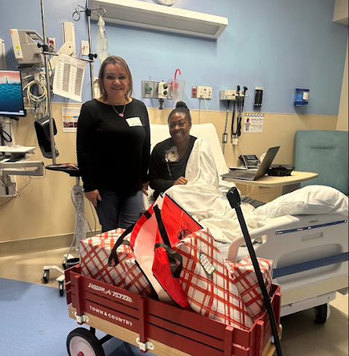 Two women in a hospital room, one sitting in bed with a white gown and the other standing beside her, smiling. A red wagon filled with checked bags and a cane is in the foreground.