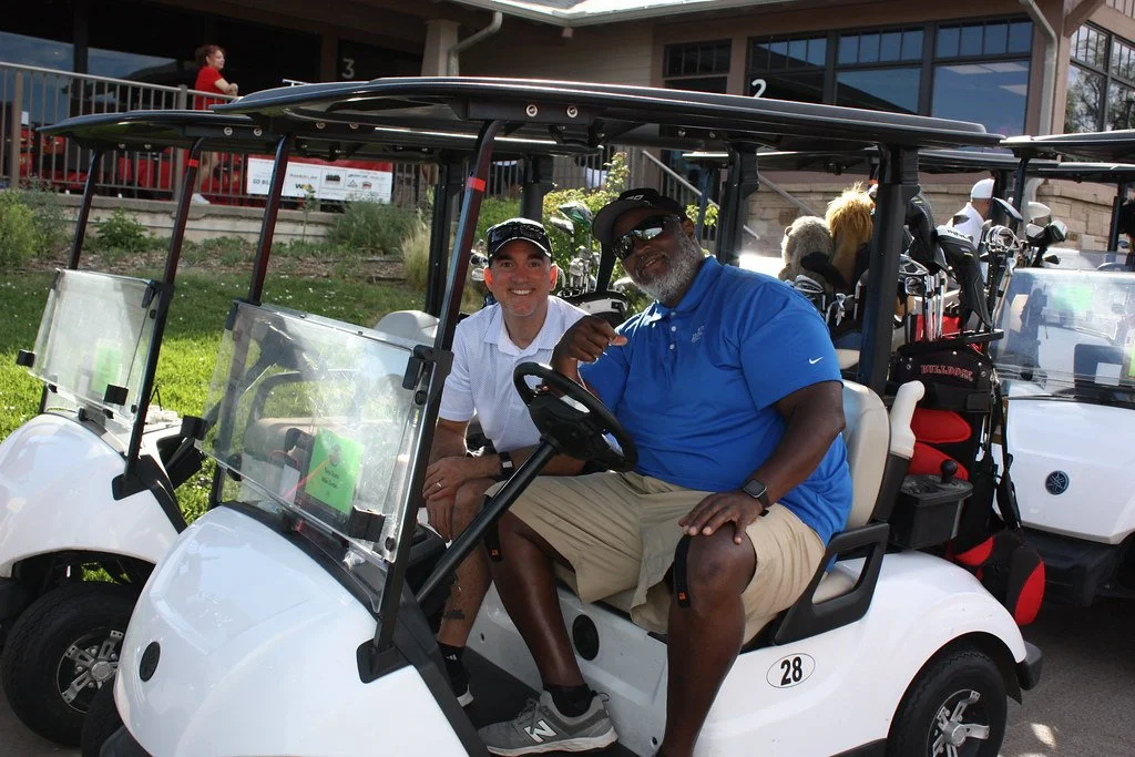 Two men sitting in a golf cart, one wearing a white polo and cap, the other dressed in a blue polo shirt, tan shorts, and sunglasses, smiling and pointing at each other. There are golf clubs in a bag behind them and other golf carts in the background.