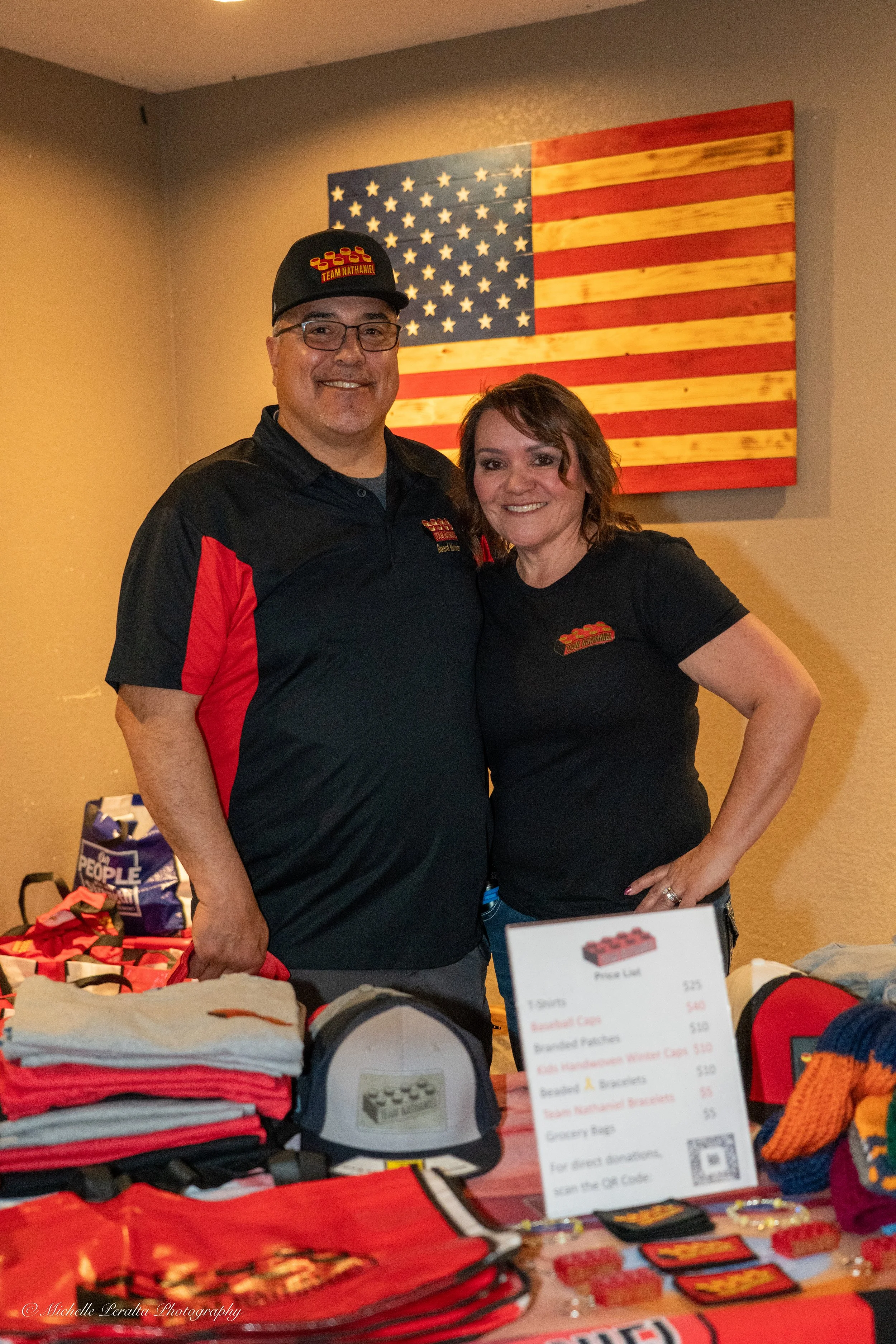 Two people standing behind a table with merchandise, smiling, with an American flag art on the wall behind them.