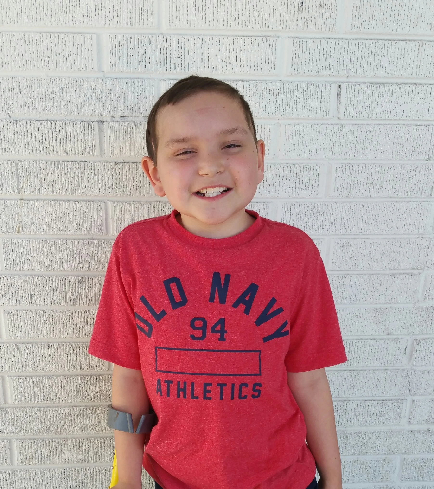 A young boy with brown hair, smiling with his eyes partially closed, wearing a red Old Navy Athletics t-shirt, standing in front of a white brick wall.