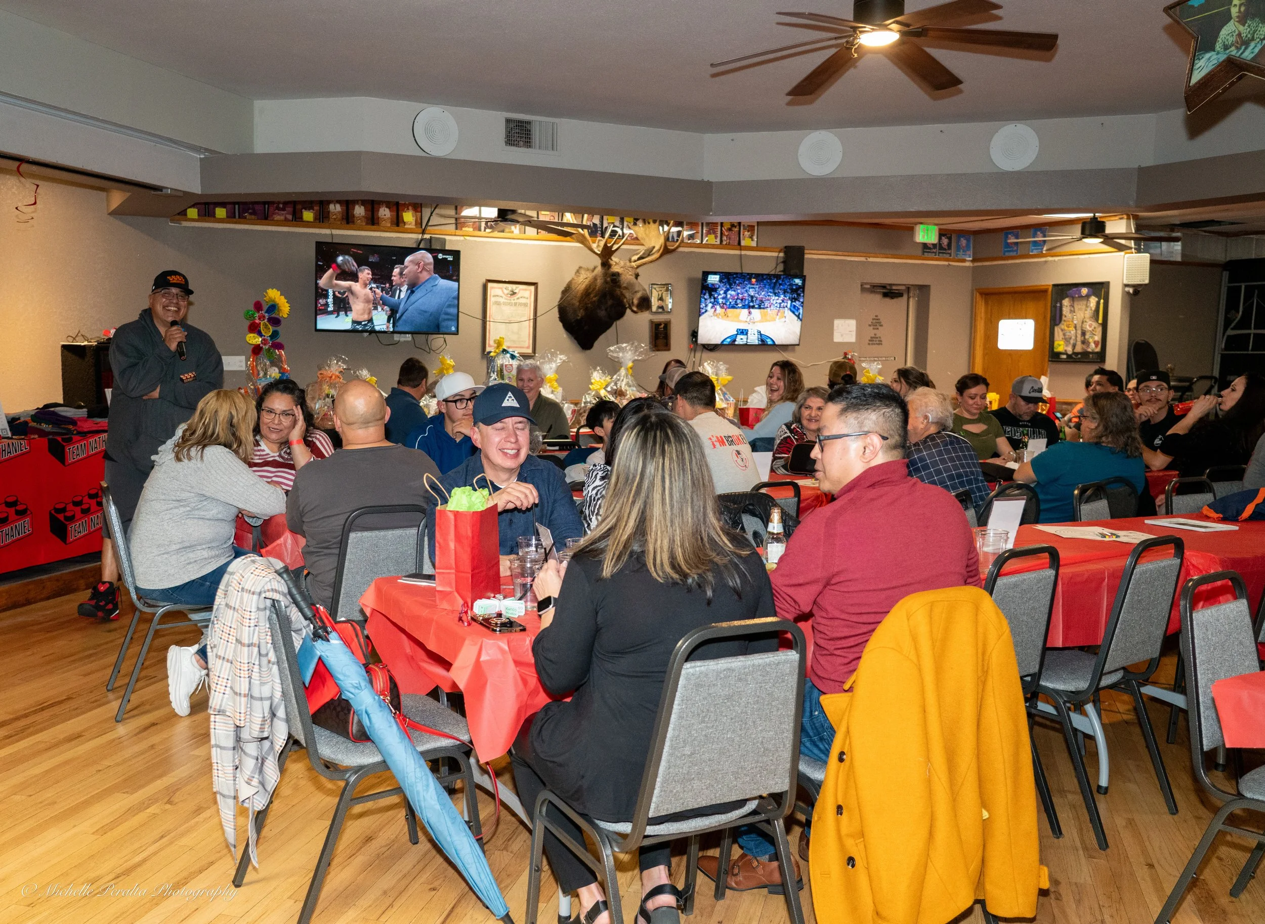 People gathered in a decorated restaurant or banquet hall, sitting at tables with red tablecloths, enjoying a celebration. A man with a microphone stands near the wall, and two large TVs display sports events. The room has wood flooring, ceiling fans, and festive decorations.