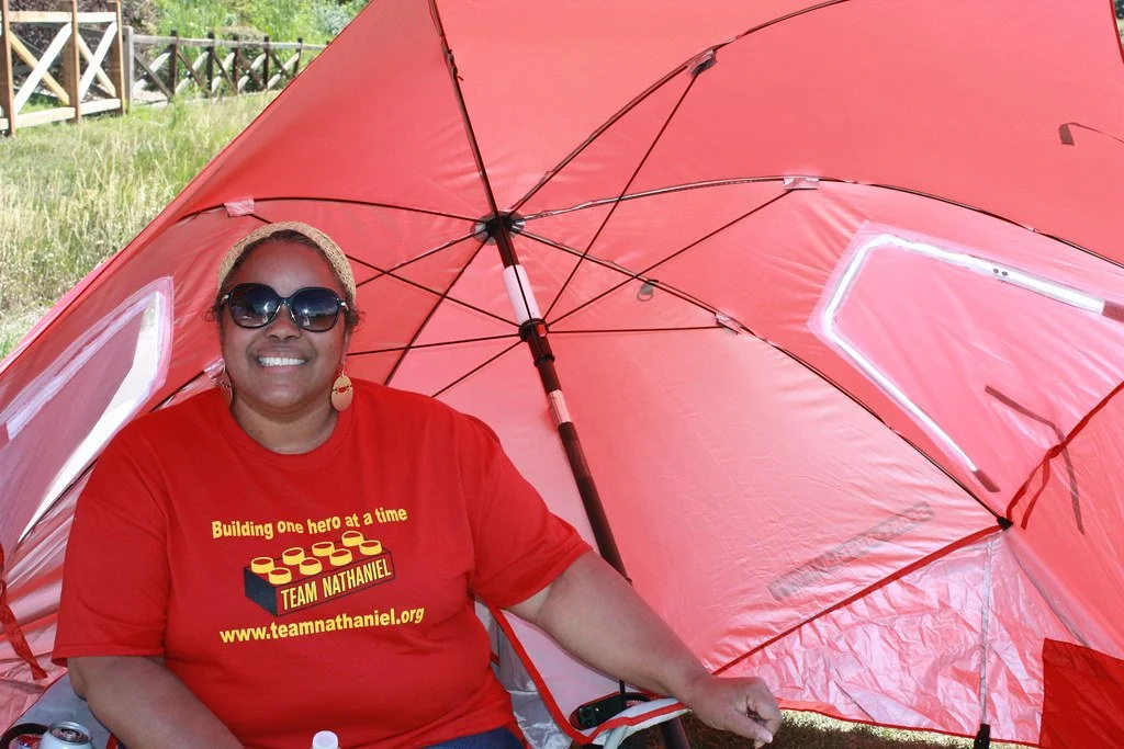 A woman smiling and posing under a large pink umbrella outdoors. She is wearing sunglasses, a beige headband, earrings, a red T-shirt with yellow and black text and graphics, and is sitting on a chair with green grass and a wooden fence visible in the background.