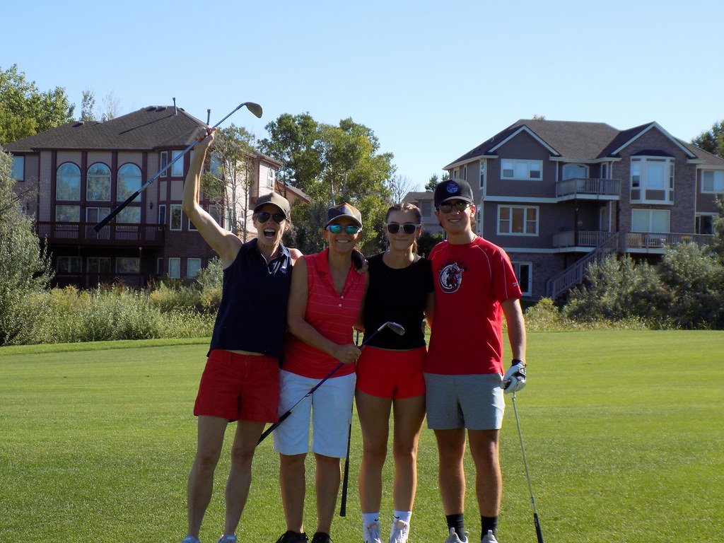 Group of four people on a golf course enjoying a sunny day, holding golf clubs, with residential buildings in the background.