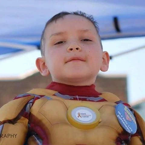 Young boy wearing a Gold Star of Hope for Cancer Awareness badge and a Disney-themed button, outdoors with blue sky and trees in the background.