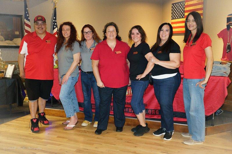 Six women standing side by side indoors, smiling, with an American flag and patriotic decorations in the background.