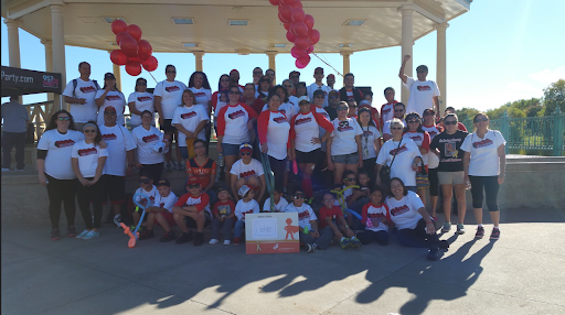 Group photograph of people wearing matching white T-shirts with red and black designs, gathered outdoors under a pavilion with red balloons hanging from the ceiling. Some children hold balloons and toys, and a small framed photo is placed on the ground in front. It appears to be a community or family event.