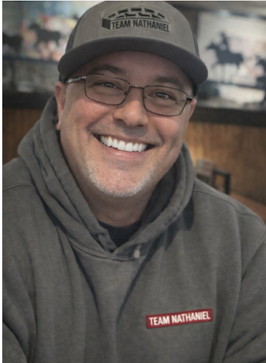 Smiling man wearing glasses, a gray hoodie, and a gray cap with a "TEAM NATHANIEL" patch, sitting indoors with a blurred background.