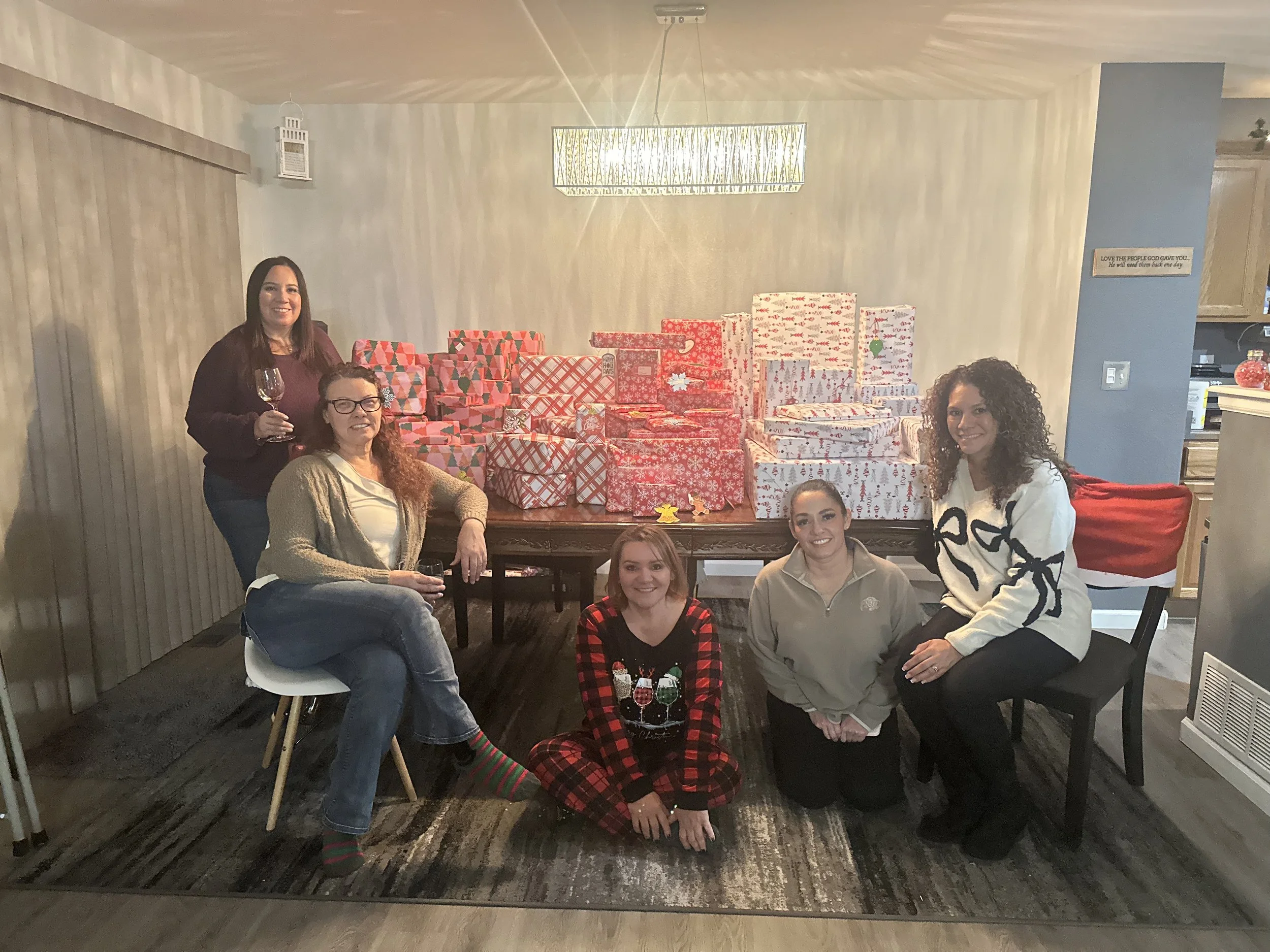 Group of five women posing in a living room with a table of wrapped Christmas gifts. One woman is standing holding a glass of wine, and the other four are seated or kneeling around the table, smiling. The background features a table filled with red and white wrapped presents, a hanging light fixture, and a mix of festive and casual decor.