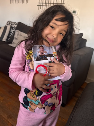Young girl with long dark hair and a pink cartoon character shirt holding a photo of herself and pointing at it in a living room.