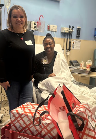 Two women in a hospital room, one sitting in a hospital bed with a blanket, and the other standing beside her. The room has medical equipment and supplies.