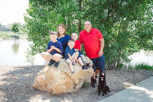 Family of five with a black dog, sitting and standing on a large rock by the river, surrounded by trees and greenery.