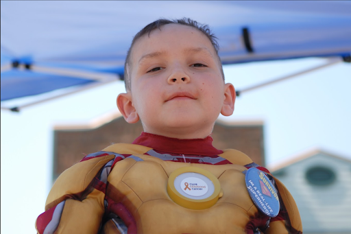 A young boy wearing a superhero costume with a yellow and red design, standing outdoors under a blue canopy.