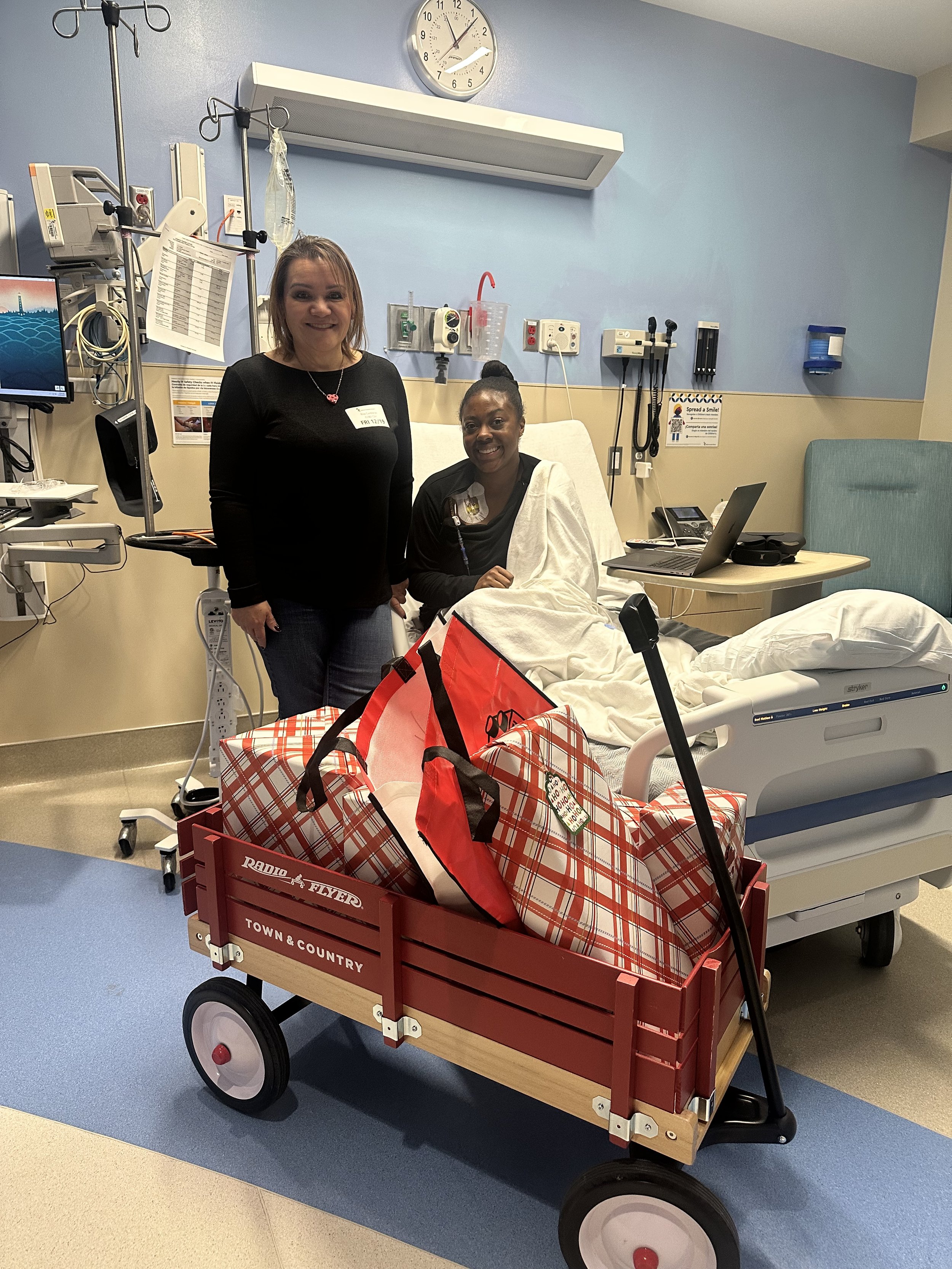 A woman in a hospital bed smiling, with two women standing beside her in a hospital room. A red wagon filled with bags is in front of the bed.