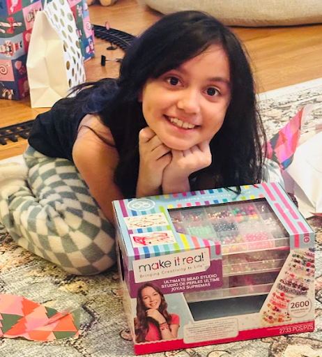 Young girl with long dark hair lying on a patterned rug, smiling and resting her chin on her hands, with a boxed craft or toy set in front of her.