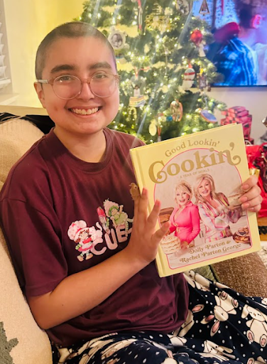 A smiling person sitting on a chair holding a cookbook titled 'Good Lookin' Cookin'' with a Christmas tree and a television in the background.