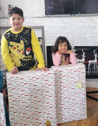 A boy and girl standing behind wrapped Christmas presents in a living room with a TV and reindeer decorations.
